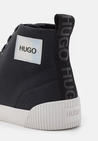 Baskets montantes noires avec semelle blanche, arborant le logo « HUGO » sur la languette du talon, une pièce latérale et le talon de la semelle dans un style minimaliste.