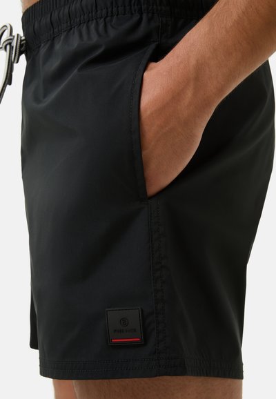 Shorts de bain noirs en matériau léger, avec une taille élastique, des poches latérales et une étiquette avec un accent rouge à l'ourlet.