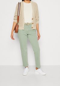 Kobieta ubrana w beżowy, dziergany sweter na białej koszuli, jasne zielone spodnie z paskiem, białe trampki i nosząca brązową torebkę na ramię.