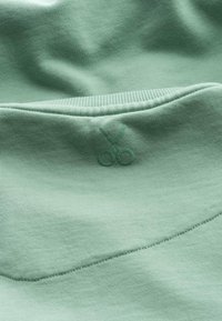 Tessuto di cotone verde chiaro con una texture liscia; presenta un design cucito con un piccolo logo di forbici ricamato con filo coordinato.