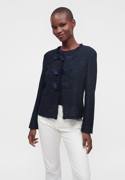 Personne debout et souriante, portant une veste bleu marine texturée avec quatre nœuds sur le devant et un pantalon blanc, sur un fond uni et clair.