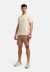 Svijetlosmeđi pamučni T-shirt s džepom na prsima; šarene kratke hlače u smeškastoj boji; bež i bijele tenisice. Jednostavan, ležeran dizajn outfita.