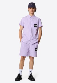 Lavendel shirt met korte mouwen en knopen, en bijpassende korte broek met zwarte logo-patches; gecombineerd met een donkere pet en zwarte schoenen.