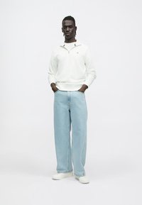 Fehér cipzáras pulóver bordás gallérral, világoskék szárszéles farmernadrág és fehér sportcipő, semleges háttér előtt állva.