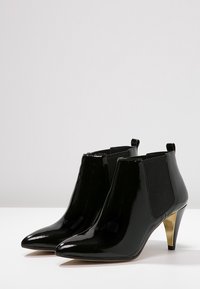 Bottines en cuir verni noir avec un bout pointu, des panneaux latéraux élastiques et un talon aiguille doré. Fini lisse avec un design épuré.