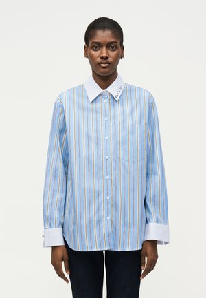 Γυναίκα που φοράει φαρδιά μπλε ριγέ πουκάμισο με λευκό γιακά και μανσέτες, στέκεται μπροστά σε ένα απλό λευκό φόντο.