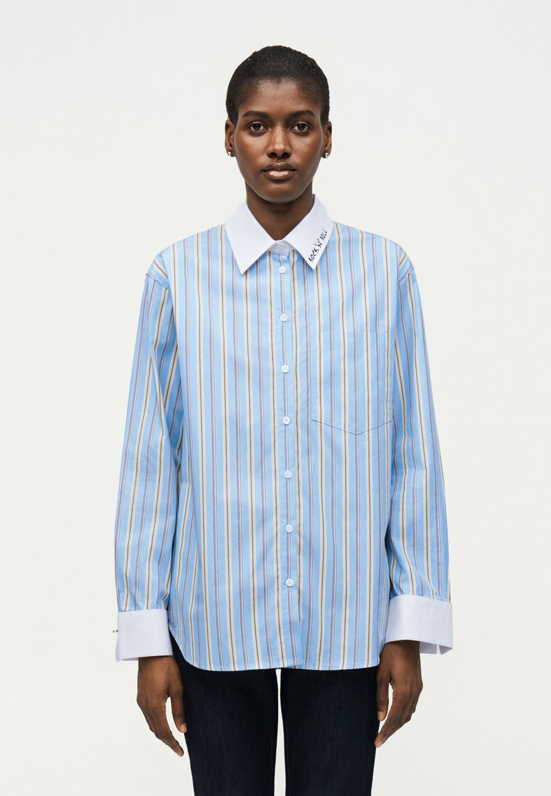 Γυναίκα που φοράει φαρδιά μπλε ριγέ πουκάμισο με λευκό γιακά και μανσέτες, στέκεται μπροστά σε ένα απλό λευκό φόντο.