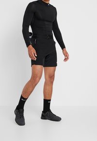 Svart långärmad sporttopp i kombination med svarta shorts som har ett bälte, bärs med svarta sportskor och strumpor. Slät textur, åtsittande design.