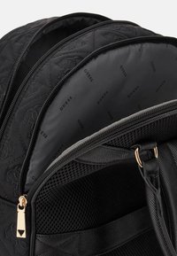 Sac à dos noir matelassé avec quincaillerie dorée, comportant deux compartiments zippés, un rangement en mesh et une doublure intérieure à motifs.