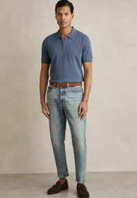 Polo cu mânecă scurtă, albastru, cu închidere prin fermoar, purtat cu blugi deschiși la culoare și curea maro. Mocasini din suede maro completează ținuta.