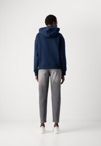 Person som har på seg en marineblå hettegenser, grå jeans og hvite sneakers, som står vendt mot en enkel hvit vegg med hendene avslappet på sidene.