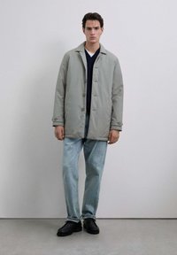 Joven con un abrigo abotonado gris claro, suéter azul marino, jeans azul claro y zapatos negros, de pie frente a una pared gris lisa.