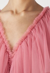 Un tissu en tulle rose tombe doucement avec des bords froncés, présentant un design en V. La texture est légère et aérienne, ajoutant une apparence délicate.