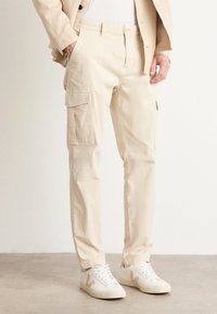 Pantalones cargo beige con bolsillos frontales y laterales, hechos de algodón, con un corte relajado y piernas ajustadas, combinados con zapatillas blancas.