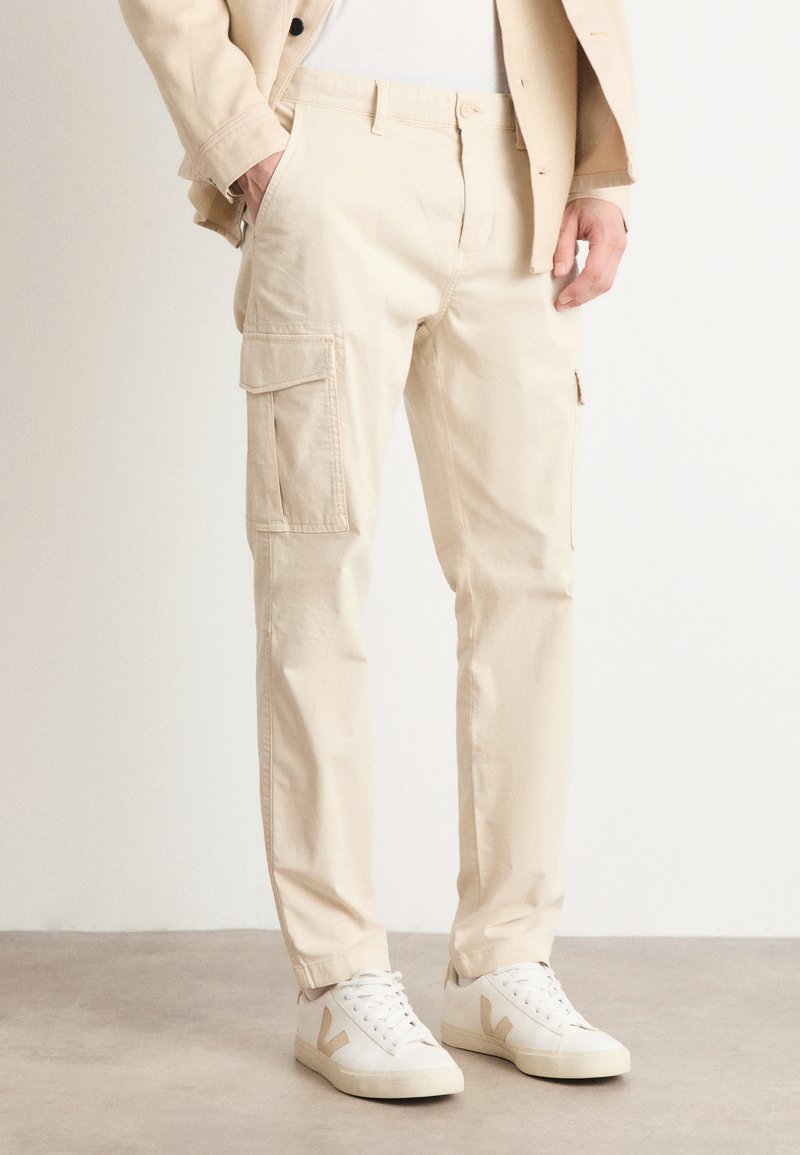 Pantalones cargo beige con bolsillos frontales y laterales, hechos de algodón, con un corte relajado y piernas ajustadas, combinados con zapatillas blancas.