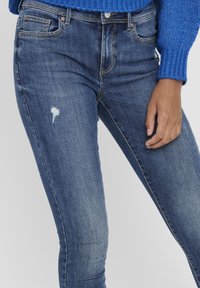 Ženska v modrih oprijetih kavbojkah z majhno raztrgano točko na levem stegnu in modrim pletenim puloverjem z rebrastimi manšetami.