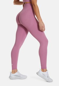 Leggings rosa a vita alta realizzati in tessuto elasticizzato, con una superficie liscia e un dettaglio con logo, abbinati a scarpe da ginnastica bianche.