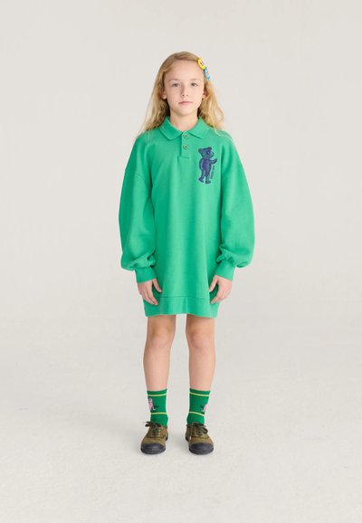 Jeune fille aux longs cheveux blonds porte une robe-sweat verte surdimensionnée avec un motif ours, des chaussettes vertes et des chaussures marron, debout devant un fond uni.