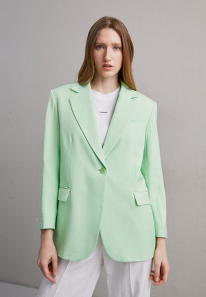 Ανοιχτό πράσινο σακάκι φτιαγμένο από ομαλό ύφασμα, με ντεκολτέ, κλείσιμο με ένα κουμπί και δύο μπροστινές τσέπες. Φορεμένο πάνω από ένα λευκό πουκάμισο.