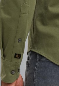 Olijfgroene blouse met lange mouwen en een knoopmanchet. Heeft een contrasterend zwart label "PINE LEGEND." De stof heeft een zachte textuur. De zoomlijn is iets gebogen.