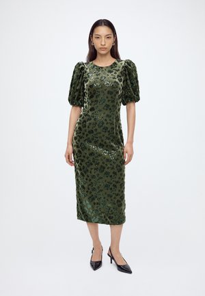 Femme portant une robe midi en velours vert avec des manches bouffantes et des talons hauts noirs, debout devant un fond blanc uni.