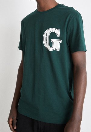 Ciemnozielona bawełniana koszulka z okrągłym dekoltem, z nadrukiem białej litery "G" i napisem "GANT" na piersi. Krótkie rękawy.