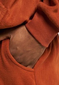 Orange fleece hoodie with a ribbed cuff. A hand is partially visible in the pocket, highlighting the soft texture and warm color.