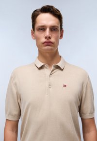 Polo beige con maniche corte, placca con due bottoni e un piccolo logo ricamato rosso e blu sul lato sinistro del petto. Tessuto morbido.