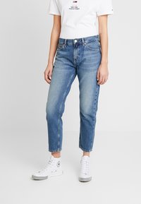 Mujer usando jeans cortos de color azul claro, zapatillas de deporte altas blancas y una camiseta blanca con un pequeño logo rojo y negro.