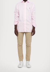 Světle růžová košile s dlouhými rukávy a knoflíky, se malým logem, v kombinaci s béžovými chinos kalhotami a bílými teniskami. Hladká bavlněná tkanina, klasický střih.