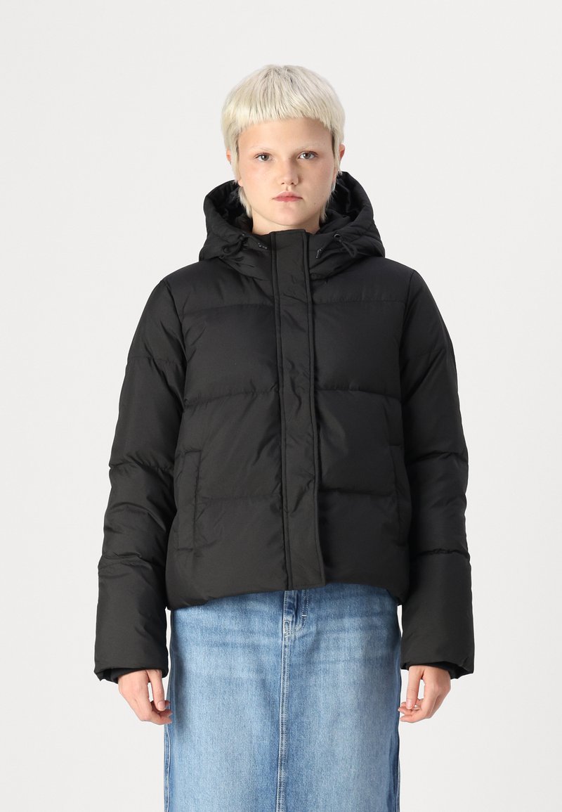 Černá péřová bunda s kapucí, prošívaný design, přední zip a elastické manžety. V kombinaci se světle modrou džínovou sukní.