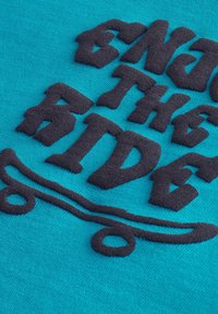 Turkos tyg med upphöjd svart text som säger "NJUT AV FÄRDET" och en skateboardgrafik, som framhäver textur och färgkontrast.