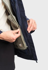 Des mains fermant la fermeture éclair d'une couche intérieure en polaire vert olive sous une veste extérieure bleu marine sur un fond uni.