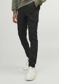Pantalons cargo noirs avec plusieurs poches, une coupe fuselée et des poignets élastiques ; portés avec des baskets blanches sur un fond neutre.