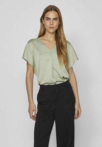 Blusa in satin verde chiaro con scollo a V e maniche corte e fluide, abbinate a pantaloni neri su misura. Texture liscia in tutto.