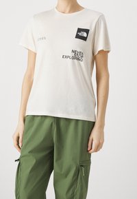 Camiseta de algodón beige claro con un gráfico negro del logo de "The North Face" y el texto impreso "NEVER STOP EXPLORING". Mangas cortas.