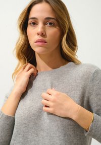 Maglione grigio chiaro in lana con scollo rotondo, polsini a costine e una consistenza morbida. Il modello tiene il collo con entrambe le mani.
