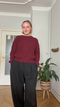 Burgundowe sweter z ribbowanym splotem i długimi rękawami, w połączeniu z czarnymi, wysokimi spodniami z plisem. Zielona roślina w plecionym koszu w pobliżu.