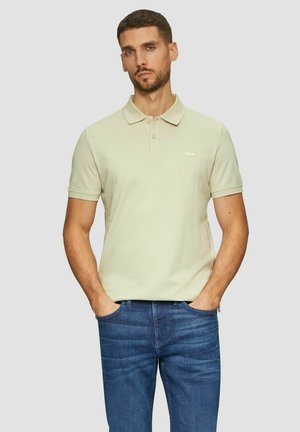 Hellgrünes Poloshirt mit Kragen und kurzen Ärmeln, versehen mit einem kleinen Logo auf der Brust, kombiniert mit blauen Jeans.