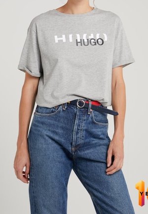 Person iført en grå T-shirt med teksten "HUGO" og højtaljede blå jeans med sort bælte mod en ensfarvet baggrund.