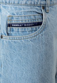 Jeans baggy de color azul claro, hechos de denim, con un bolsillo frontal, costuras visibles y una etiqueta de tela oscura con la marca "REELL BAGGY".