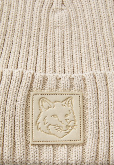 Cream-colored knit beanie with vertical ribbing, featuring a textured beige leather patch with an embossed fox design at the front.