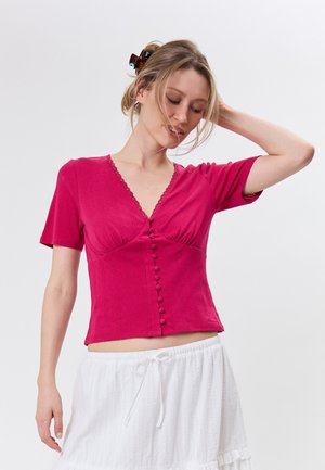 Kobieta o blond włosach w spince do włosów, ubrana w krótki magentowy top z guzikami i białą spódnicę, pozująca z jedną ręką uniesioną za głową.