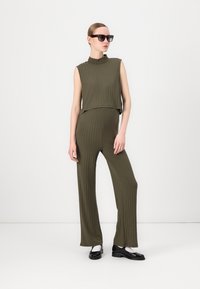 Conjunto din două piese, de culoare verde măsliniu, cu un top fără mâneci și pantaloni evazați. Modelul poartă ochelari de soare negri supradimensionați și pantofi negri.