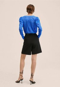Blouse bleue à pois avec manches bouffantes, associée à un short noir coupé sur mesure et des talons noirs à lanières, vue de dos.
