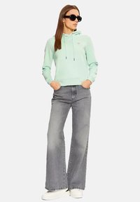 Felpa verde menta con cappuccio e logo, abbinata a jeans grigi a vita alta e gamba larga e scarpe da ginnastica bianche. Accessoriata con occhiali da sole neri.