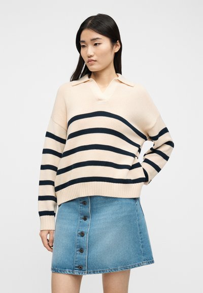Νεαρή γυναίκα που φοράει μπεζ πουλόβερ με μαύρες οριζόντιες ρίγες και μπλε τζιν φούστα με κουμπιά μπροστά, στέκεται μπροστά σε απλό φόντο.