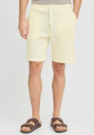 Shorts de couleur jaune clair à texture en gaufre, avec une taille élastique et un cordon de serrage, dotés de poches latérales, associés à des sandales à glissière marron.