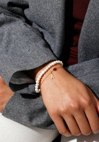 Bracelet with white pearls and a delicate gold chain featuring a red heart and a gold letter 'A.' Hand rests on a grey fabric.
