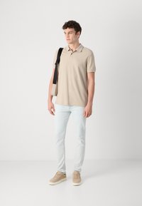 Svijetlosmeđi polo majica, slim-fit svijetloplave traperice i bež tenisice. Crna torba za rame na ramenu, model stoji na neutralnoj pozadini.